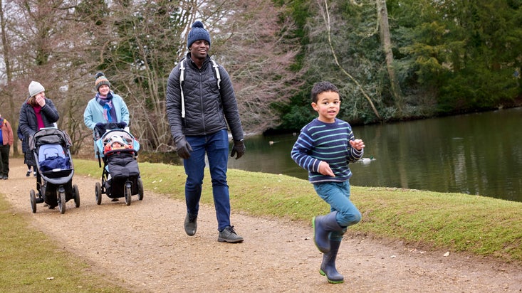 A family taking a walk around the parkland at Stowe in Buckinghamshire. Two women with pushchairs are seen on the path in the background while a young child and a man lead ahead of them. A lake can be seen behind them.